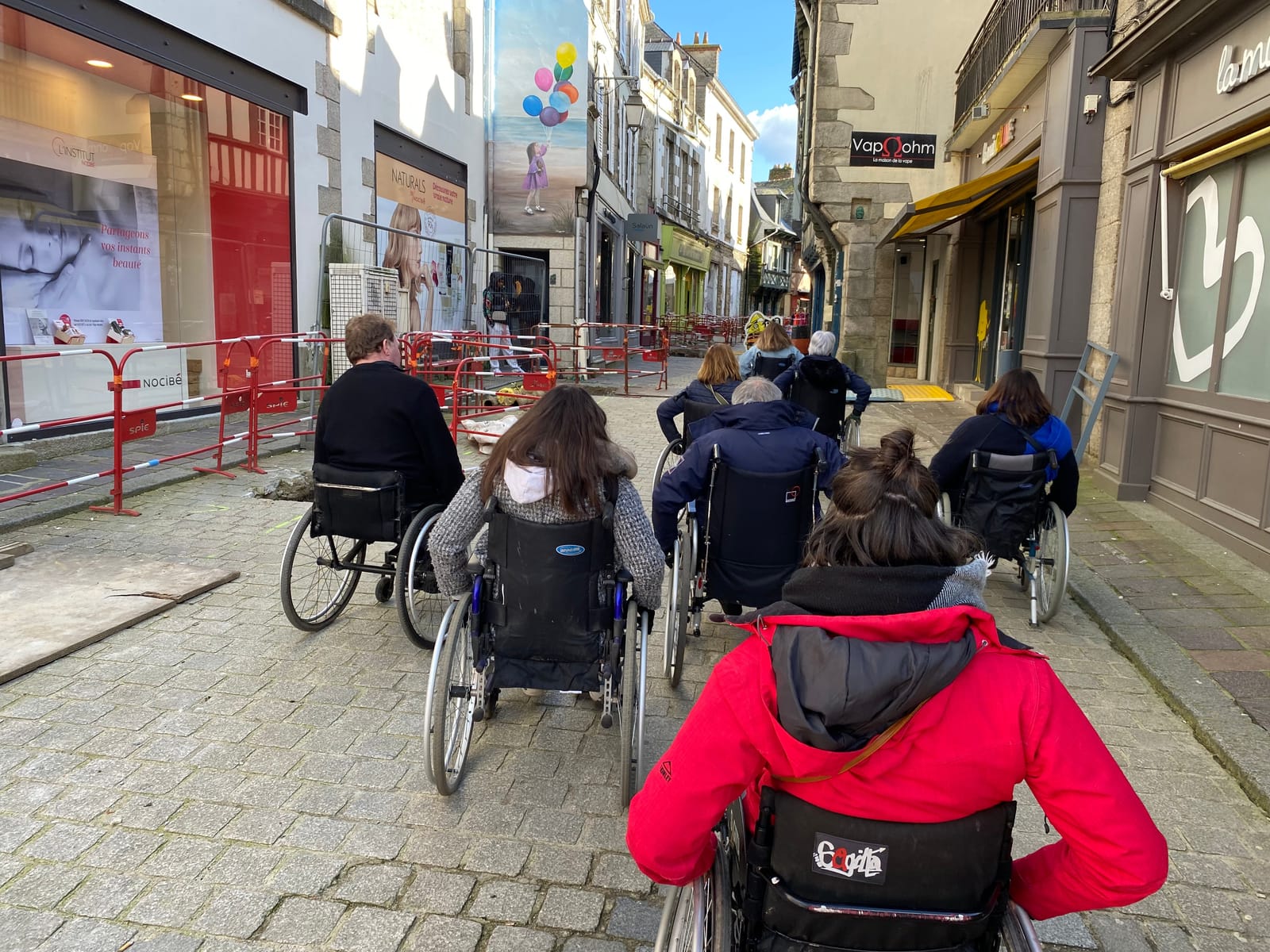 8 personnes de dos en fauteuils roulant dans une rue qui est en travaux. Le trottoir est bloqué par des barrières et les pers